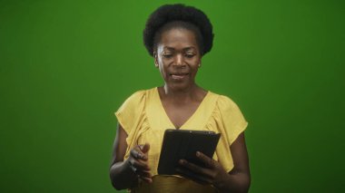 Yeşil ekran stüdyosunda elinde tablet tutan, gülümseyen ve tıklayan siyah kadın ayakta ve içeriği okurken; sakin odaklanma.