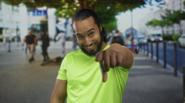 Neon yeşil tişörtlü adam gülümseyen ve işaret eden kamera meşgul şehir caddesinde; dostça meydan okuma oyuncu.