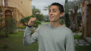 Man in grey sweater winks eye and points thumb at historic old town building courtyard outdoors; confidence.