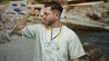 Genç adam, deniz kenarında steteskop takarken kaslarını kasıyor. Gün içinde canlı bir plaj zeminine karşı dayanıklılık ve sağlığını somutlaştırıyor..