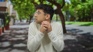 Krem rengi süveter giyen ve yuvarlak gözlükler takan bir adam, yeşil ağaçların altında göğsüne yapışmış eller, baş aşağı dönüyor, endişe özlemi.