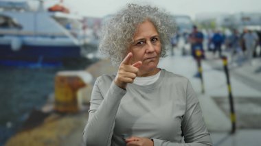 Gri saçlı bir kadın deniz kıyısında durup düşünceli bir jest yapıyor ve açık deniz ortamının ortasında düşünceli bir ifade sergiliyor..