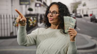 Şehirdeki kadın Amerikan dolarlarını elinde tutuyor. Gözlük ve beyaz süveter takıyor. Açık hava ortamında başarıyı ve orta yaşlı cazibesini somutlaştırıyor..