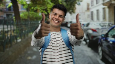 Şehir caddesinde başparmağıyla gülümseyen genç bir adam, çizgili bir gömlek ve mavi bir sırt çantası, park etmiş arabalar ve ağaçlarla çerçevelenmiş, dışarıda pozitiflik yayıyor..