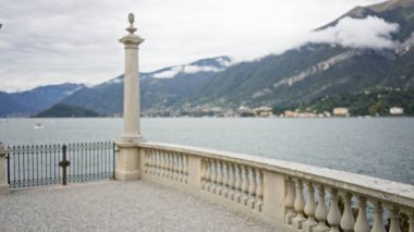 Odaklanamadığı su ve uzak dağları olan bir gölün kıyısındaki taş balystrade terası, yumuşak bokeh açıklığı; arkaplan telif alanı sakin.