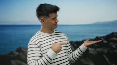 Çizgili tişörtlü genç İspanyol bir adam kumsalda durmuş avucunu işaret ediyor ve sakin bir deniz manzarasına başparmak işareti yapıyor.