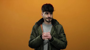 Yeşil ceketli genç İspanyol bir adam izole edilmiş turuncu bir arka planda dikiliyor. Yüzünde düşünceli bir ifadeyle serin bir ifade ve sımsıkı tutulmuş eller soğuk havayı işaret ediyor..