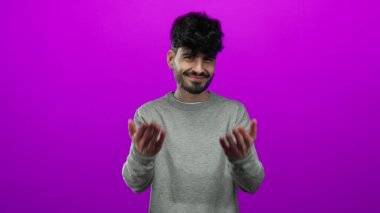 Genç İspanyol bir adam gülümsüyor ve elleri ile işaret ediyor. İzole edilmiş, pembe bir arka planı var. Gri bir süveter giyiyor.