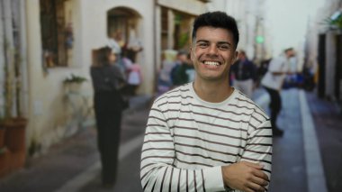Şehir caddesinde gülen yakışıklı genç bir adam çizgili bir tişört giyiyor ve arka planda insanlar canlı bir şehir manzarası yansıtıyor..