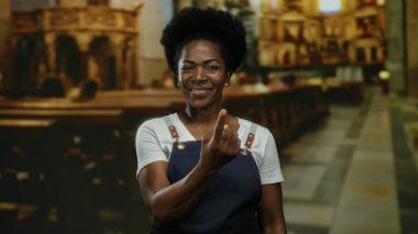 Önlüklü kadın kilise ortamında parmaklarıyla jest yapıyor, geleneği ve çağdaş stili ev ortamında güven ve cazibeyle harmanlıyor..