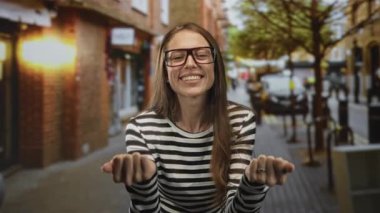 Gözlüklü bir kadın kalabalık bir şehir caddesinde iki eliyle işaret eder, geniş bir gülümseme; günlük eğlence karşılama.