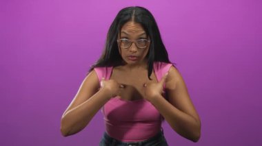 Stüdyoda pembe askılı ve mor arka planda gözlük takan kadın işaret parmağıyla göğsünü işaret ediyor..