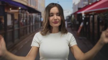 Genç bir kadın, gün ışığında dar bir sokakta iki eliyle kameraya dönük tırnak işareti yapıyor. Kafe tenteleri görünür durumda..