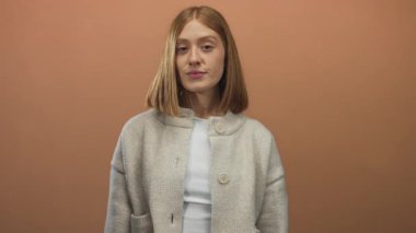 Genç sarışın kadın düz kahverengi duvarlı parlak stüdyoda sağ eliyle gözlerini kapar; neşeli sürpriz neşe.