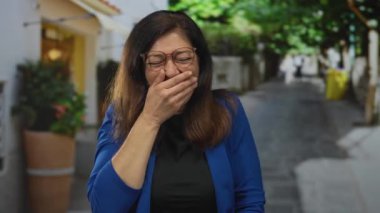 Gözlüklü ve mavi ceketli bir kadın, güneşli bir şehir caddesinde gülerken ağzını kapatır..