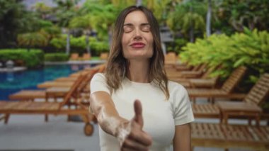 Beyaz tişört giyen bir kadın, açık havuz kenarında başparmağıyla bir jest gösteriyor..