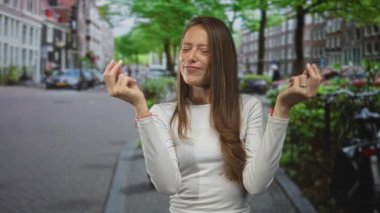 Kadın, bir şehir caddesinde iki eliyle çimdikleme hareketi yapıyor, beyaz gömlek ve bilezik görünür; oyuncu güven.