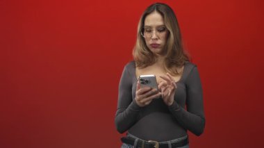 Akıllı telefon tutan ve kırmızı stüdyo arka planında çapraz kollu gözlük takan kadın, kot kemeri görünür; sessiz tefekkür.
