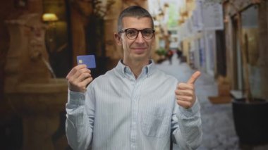Gözlüklü beyaz bir adam kredi kartı tutuyor ve bulanık bir geçmişi olan Avrupa 'nın pitoresk caddesinde güvenle el sallıyor..