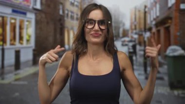 Kadın her iki tarafı da işaret ediyor, kolsuz tişört ve geniş yuvarlak gözlük takıyor, görünür omuzlar ve göğüs, neşeli sırıtış; oyuncu merak.