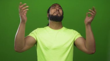 Stüdyoda, neon bir tişört giyerken elini sıkan ve yüzünü yukarı kaldıran bir adam, hayal kırıklığını savunan bir mücadele içindedir..