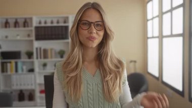 Gözlüklü sarışın kadın, ofis ortamında elleri masasında çenesini çerçeveliyor..