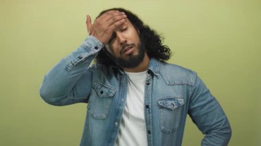 Açık yeşil bir duvara karşı stüdyoda eli alnında ve görünür sakallı bir adam; yorgunluk yansıması.