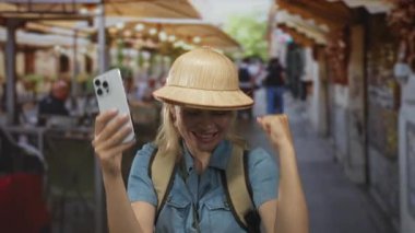 Saman başlık ve sırt çantasıyla akıllı telefon tutan ve terasta yumruğunu kaldıran kadın, geniş bir gülümsemeyle; neşe.