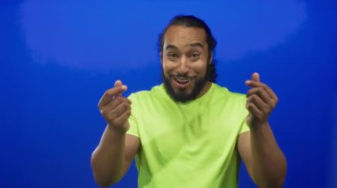 Mavi bir stüdyoda, neon yeşil gömlekli bir adam parmaklarını çimdiklerken gülümsüyor..