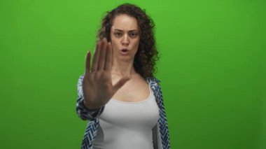 Avuç içleri açık genç bir İspanyol kadın yeşil stüdyoda, beyaz atlet bluz ve desenli gömlek giyerek durmak için el hareketi yapıyor..