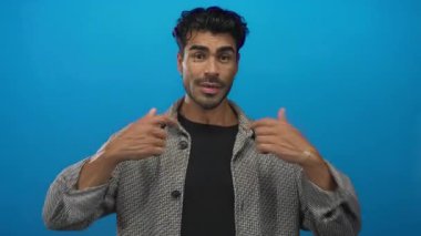 Gri ceketli adam mavi stüdyoda parmaklarını göğsüne işaret ediyor..