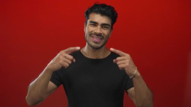 Siyah tişört giyen genç İspanyol adam kırmızı stüdyo ışıkları altında geniş bir gülümsemeyi işaret ediyor..