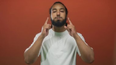 Sakallı bir adam stüdyoda parmaklarını turuncu arka planla yanaklara bastırıyor, beyaz tişört giyiyor ve ön tarafa bakıyor;.