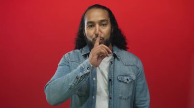 Sessiz kalmak için parmakları dudaklarına kadar uzanan adam, kot ceket giyiyor ve kırmızı stüdyoda görünen sakallı beyaz tişört giyiyor; gizli sessizlik.