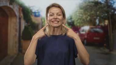 Sarışın kadın, taştan duvarlar ve park halindeki kırmızı bir arabayla güneşli bir sokakta yanakları işaret ediyor..