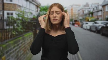 Sarı saçlı genç bir kadın şehir caddesinde yüzünü düzeltiyor. Etrafında arabalar ve binalar var. Şehir caddesinde gün ışığında siyah bir bluz giyiyor..