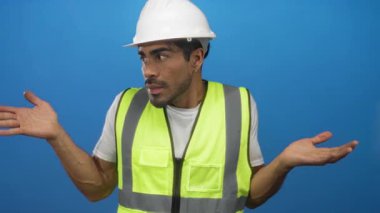 Çağdaş stüdyoda çelik yelek ve sert şapkalı bir adam avuçlarını mavi zemine yaslıyor; belirsizlik.