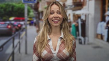 Gündüz vakti işlek bir şehir caddesinde gülümserken, uzun saçlı sarışın kadın parmağıyla başını işaret ediyor..