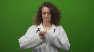 Steteskoplu beyaz laboratuvar önlüklü genç İspanyol kadın doktor, yeşil stüdyoda göğsüne yakın parmakları kenetliyor; belirsizlik endişesi.