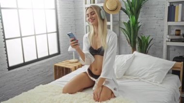 İç çamaşırı ve bornoz giyen kadın, stüdyoda diz çökerken akıllı telefon tutuyor ve pencereden güneş ışığı süzülüyor..