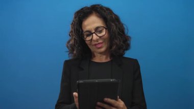 Mavi arka planda tablet kullanan, siyah takım elbiseli, güvenle gülümseyen, profesyonel ve teknolojik konuları vurgulayan İspanyol bir kadın..