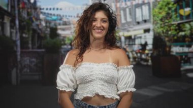 Bir şehir festivalinde elleri bağlı ve çıplak omuzlarıyla gülümseyen kadın, beyaz bluz ve kolye giyiyor; sevinç güveni iyimserliği.