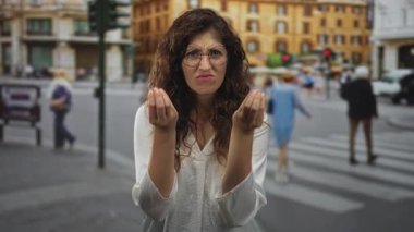 Avuçları havada ve elleri sorgulayan bir kadın, kalabalık bir şehir yaya geçidinde, kaşlarını çatarak ve sessizce sorarken öne eğilerek hareket eder;.
