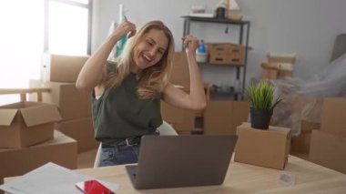 Yeni döşenmiş oturma odasında kutular ve saksı bitkileriyle dizüstü bilgisayar kullanarak neşeli ve heyecan verici bir atmosfer yaratarak mutlu bir kutlama yapan kadın..