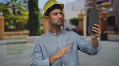 Açık hava parkı ortamında tablet kullanan İspanyol asıllı genç adam, inşaat ortamında yeşil zemin ile modern teknoloji kullanımını gösteriyor..
