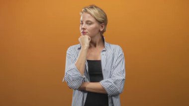 Katlanmış kolları ile çene üzerinde dinlenen kadın, siyah üzerine çizgili gömlek giyiyor turuncu bir stüdyo arka planı karşı, sabit bakışlar; düşünceli.