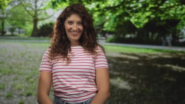 Kadın, çizgili tişört ve yeşil ağaçların altında açık ön kol ile parkta sağ parkı işaret ediyor; keyif daveti.