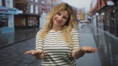 Çizgili gömlekli bir kadın ıslak bir kaldırım taşı sokakta avuçlarını açık tutarak dükkanlar ve binalar arkasında; neşeli karşılama.