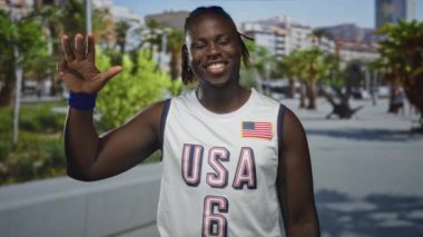 Genç adam beyaz ABD forması ve bileklik giymiş el sallıyor, yükseltilmiş avuç içi ve gülümseme gösteriyor; dostça selamlaşma.