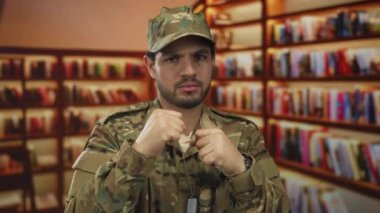 Kamuflaj üniformalı genç İspanyol asker kitaplıklarla çevrili bir kütüphane binasında göğüs hizasında yumruklarını kaldırıyor..